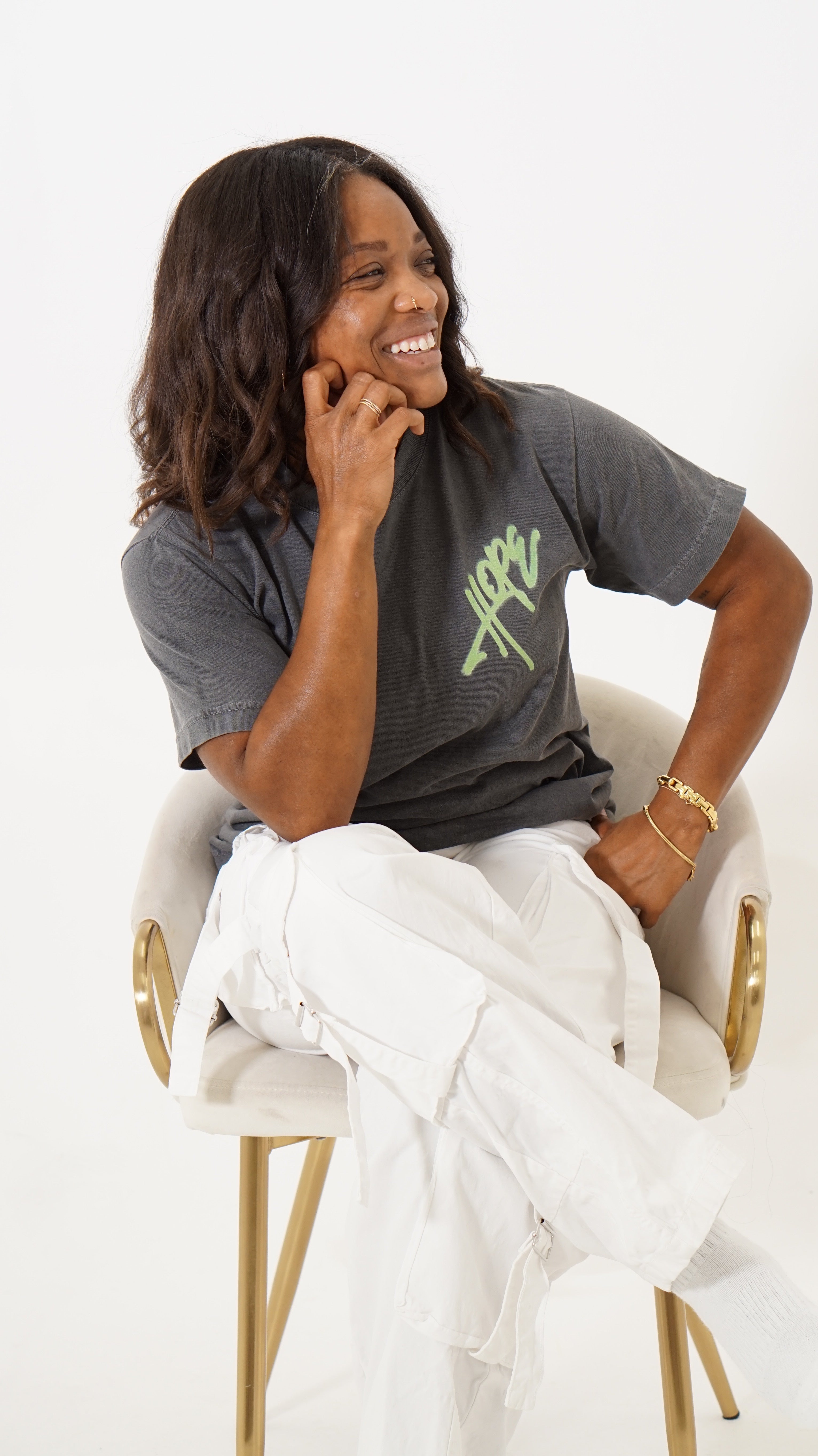 A woman sitting on a white chair with gold legs, wearing a dark gray t-shirt with a green graphic and white pants, against a plain white background.